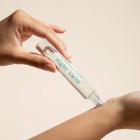 Hand applying a skincare product labeled 'Night Swim' to a wrist against a beige background