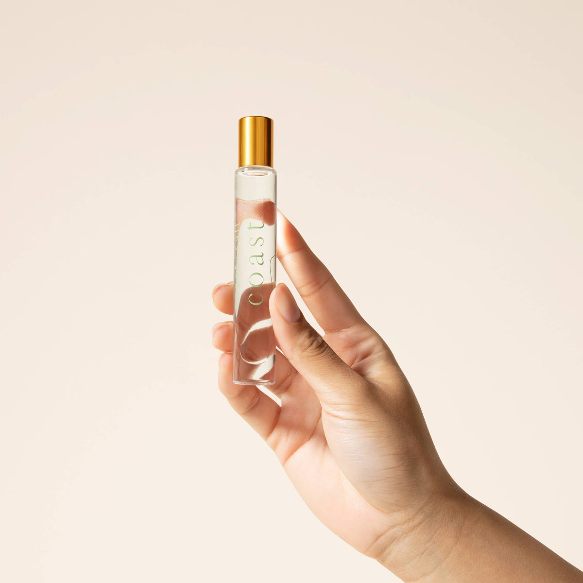 Hand holding a clear perfume bottle with a gold cap against a beige background