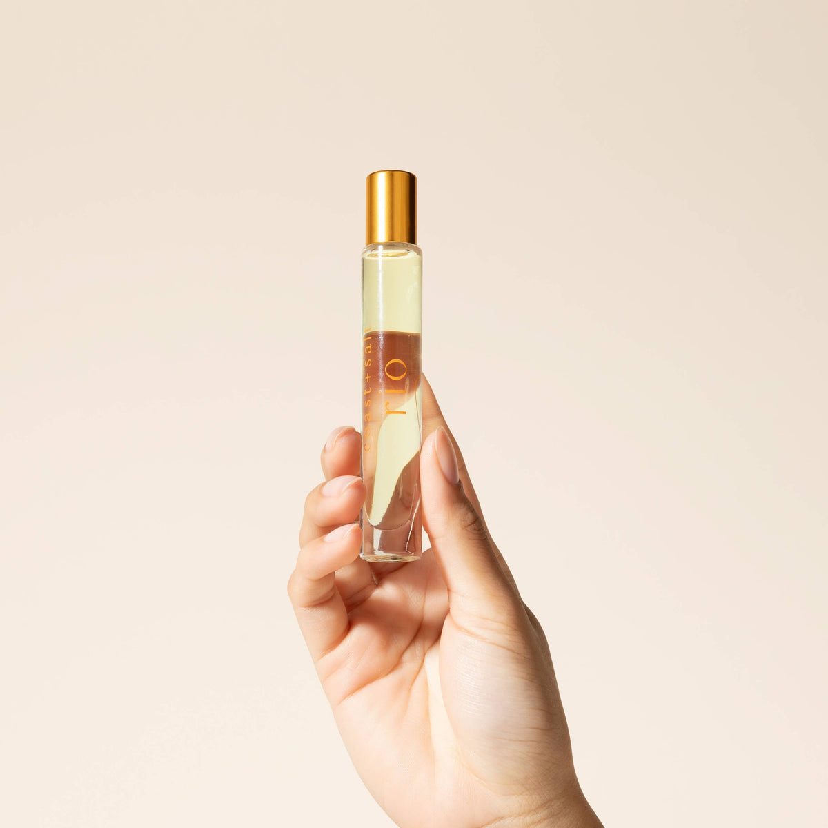 Hand holding a small perfume bottle with a gold cap against a beige background