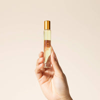 Hand holding a small perfume bottle with a gold cap against a beige background