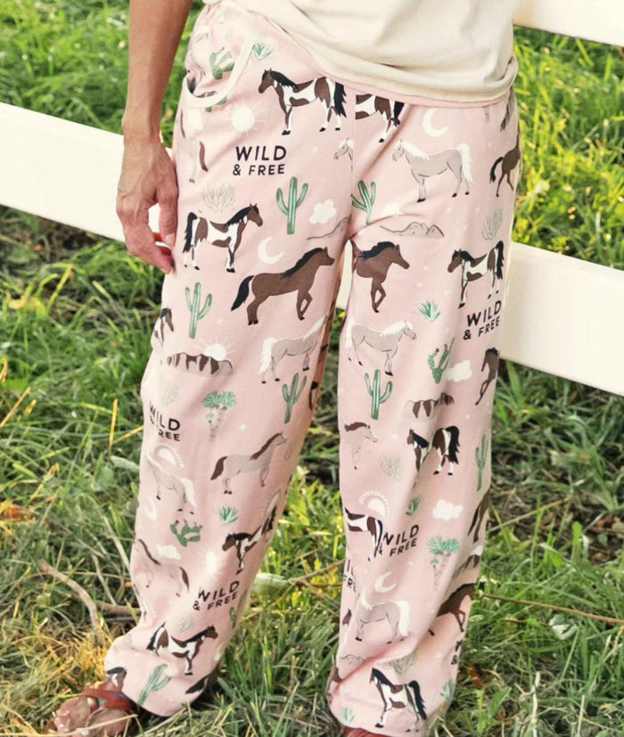 A pair of pink women's pajama pants with a horse and desert pattern, featuring text that reads 'WILD & FREE'.