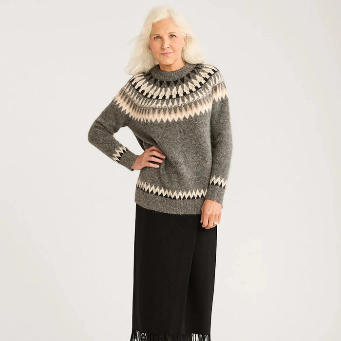 A woman wearing an oversized alpaca fair isle sweater with geometric patterns, predominantly gray, and black skirt with fringe detailing.
