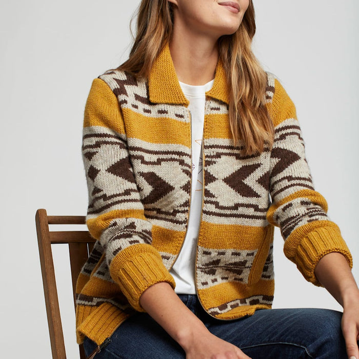 A woman sitting and wearing a zip-front cardigan with geometric patterns in yellow, brown, and white colors.