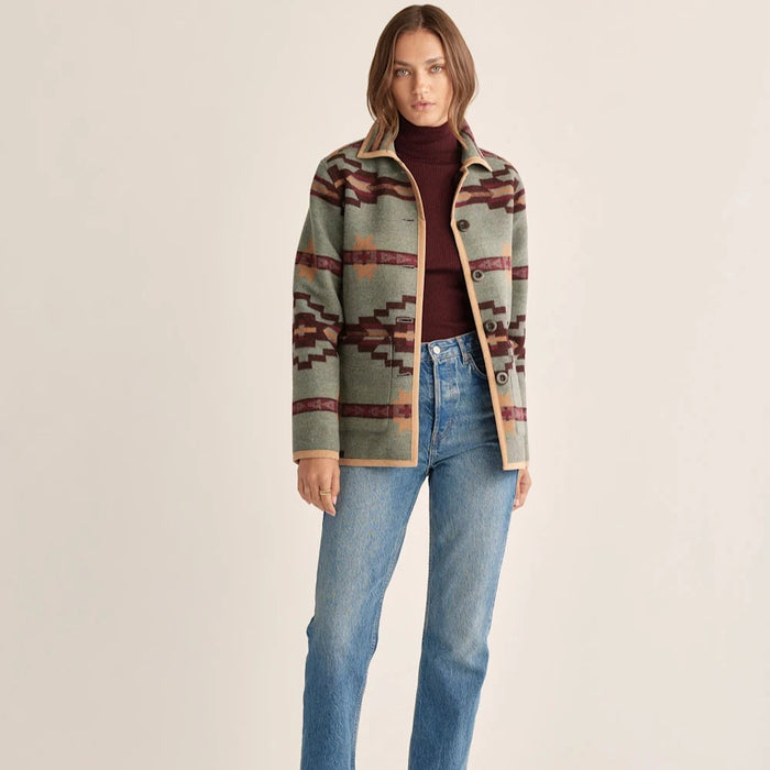 A woman models a Pendleton reversible barn coat with a geometric pattern in beige, red, and green colors over a red top, paired with blue jeans and brown boots.