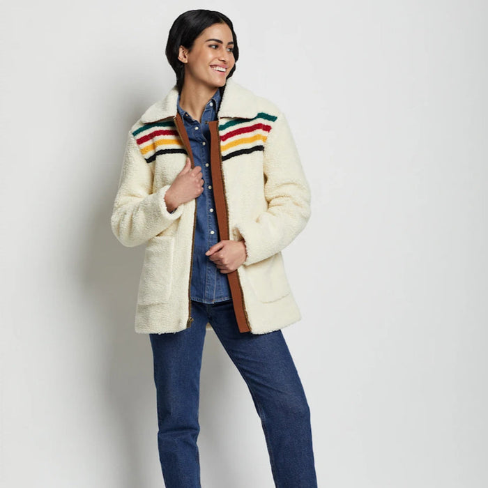 A woman models a beige Sherpa fleece jacket with colorful stripes on the collar, paired with blue jeans and tan boots.