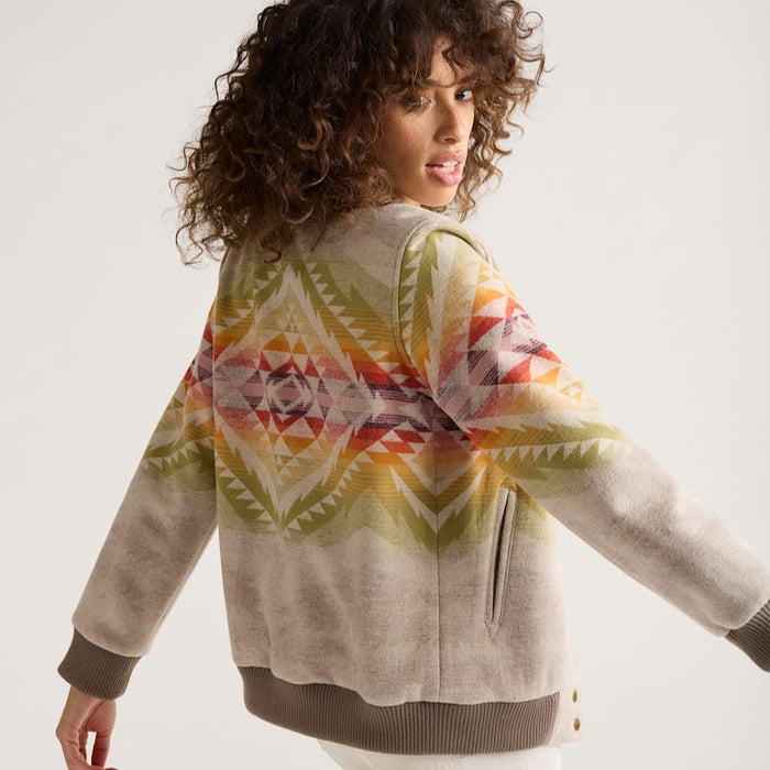 A woman is shown from the back, wearing a beige, multicolored geometric patterned wool bomber jacket with a snap front, knit ribbing at the neck, cuffs, and hem.