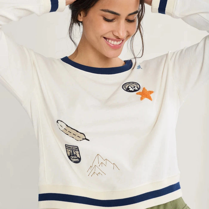 A woman wearing a white long-sleeved crewneck sweatshirt with embroidered graphics and Pendelton branded patches, paired with green pants.